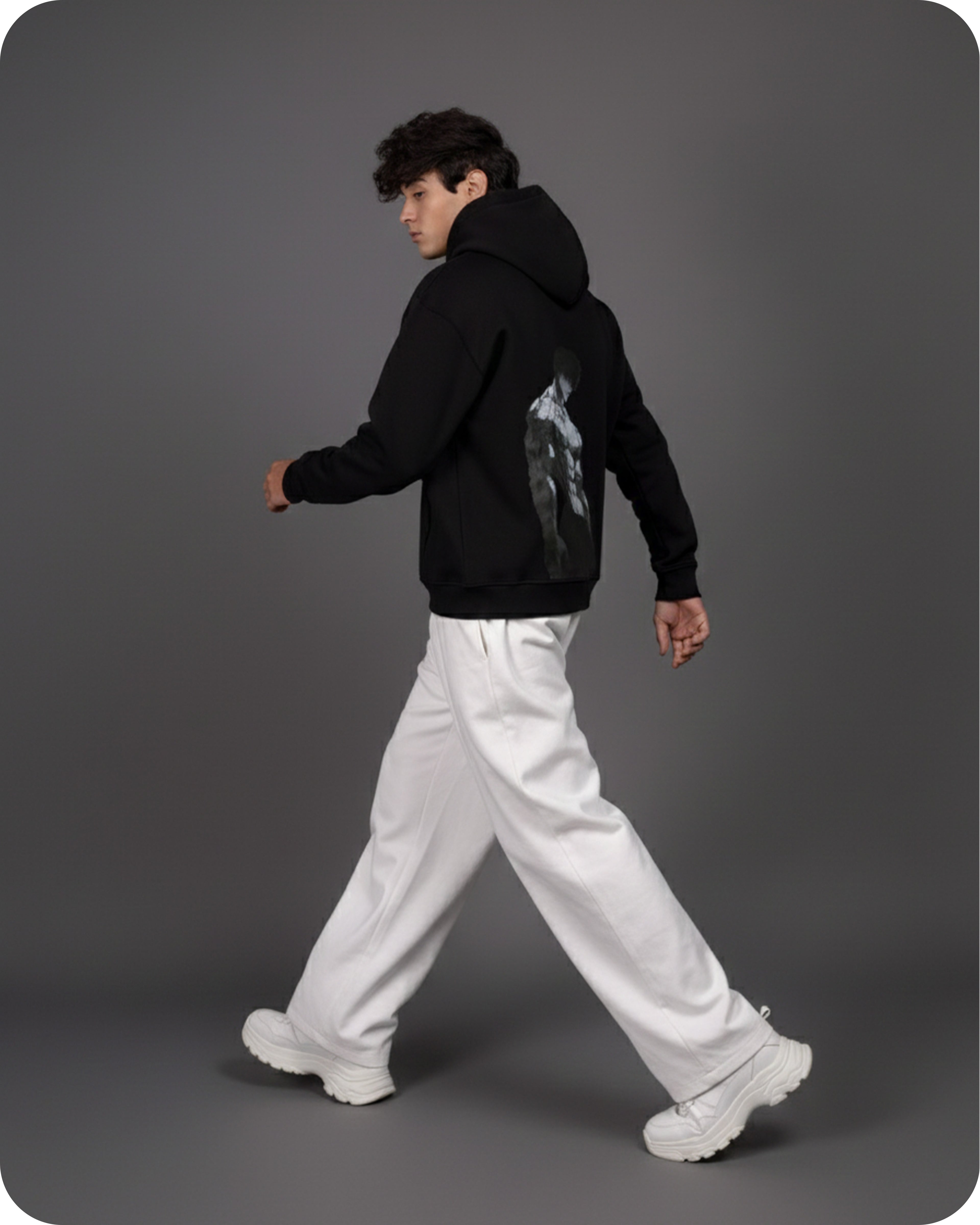 Person wearing a black hoodie with a graphic design, white pants, and white sneakers on a gray background