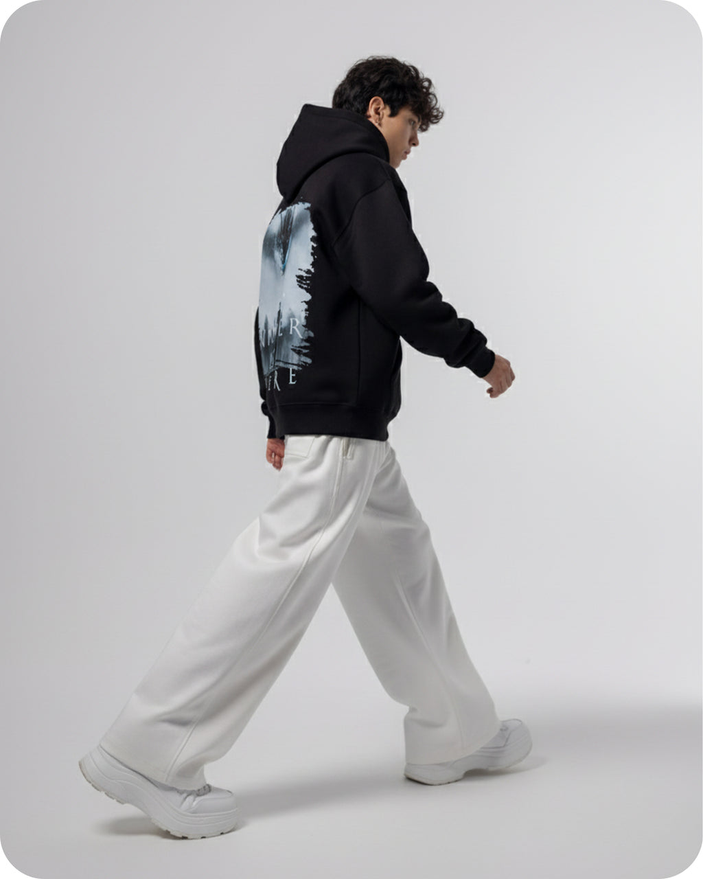 Person wearing a black hoodie with a nature graphic and white pants on a light gray background