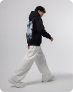 Person wearing a black hoodie with a nature graphic and white pants on a light gray background